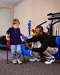 Imagem do menino novo, com bastões, durante uma sessão de fisioterapia